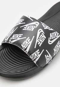 Černé skluzné sandály se širokým páskem pokrytým opakujícími se bílými logy "NIKE". Texturovaná černá stélka s drážkami pro lepší přilnavost.