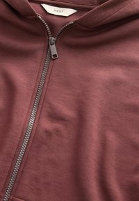 Bordeauxfarbener Zip-Hoodie aus weichem Material. Verfügt über einen Metallreißverschluss mit einem Plastik-Ziehband und einem Etikett mit der Aufschrift "NEXT" im Kragen.