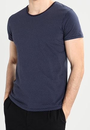 Mann trägt ein marineblaues, kurzärmeliges, gemustertes T-Shirt mit den Händen in den Taschen einer schwarzen Hose vor einem einfarbigen Hintergrund.