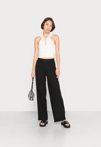 Top halter branco com textura em nervuras e frente com cordas, combinado com calças largas pretas. A modelo segura uma bolsa listrada preta e branca, usando sandálias pretas.