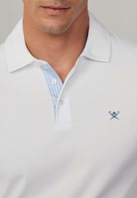 Weißes Poloshirt mit einer hellblauen gestreiften Knopfleiste, zwei Knöpfen und einem kleinen blauen gestickten Detail auf der Brust. Glattes Baumwollmaterial.