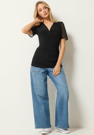 Mujer de cabello rubio con un top negro de estilo cruzado con mangas cortas transparentes, jeans azules de pierna ancha y zapatillas blancas, de pie contra una pared clara.