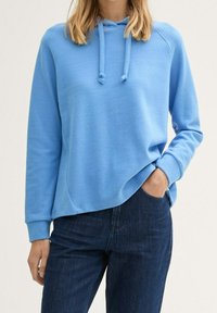 Ljusblå huvtröja med en avslappnad passform, dragsko och ribbade ärmslut, tillsammans med mörka denimjeans. Slät tygtextur.