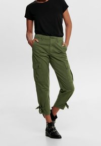 Groene cargobroek met een relaxed fit, voorzien van zijzakken, strikdetails bij de boorden en een klassieke tailleband, gecombineerd met een zwart t-shirt.