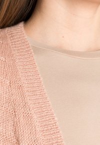 Ljusrosa stickad kofta med ett texturerat stickmönster, lager på lager över en slät beige åtsittande t-shirt, som visar tygets mjukhet.