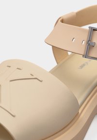 Μπεζ σανδάλι με ανοιχτά δάχτυλα, με ανάγλυφο λογότυπο «K» στο μπροστινό λουράκι και ρυθμιζόμενο ασημένιο λουράκι με αγκράφα στον αστράγαλο.