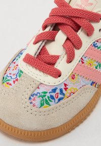 Adidas shoes featuring pink fabric laces, a cream leather upper, suede toe cap, and colorful floral patterns on a blue background.