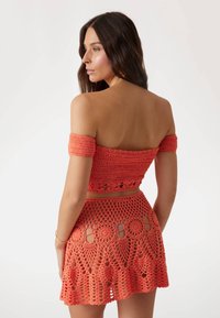 Gehaakt tweedelige set in helder koraal, bestaande uit een off-the-shoulder top en een rok met ronde uitsparingen en textuurs patronen.