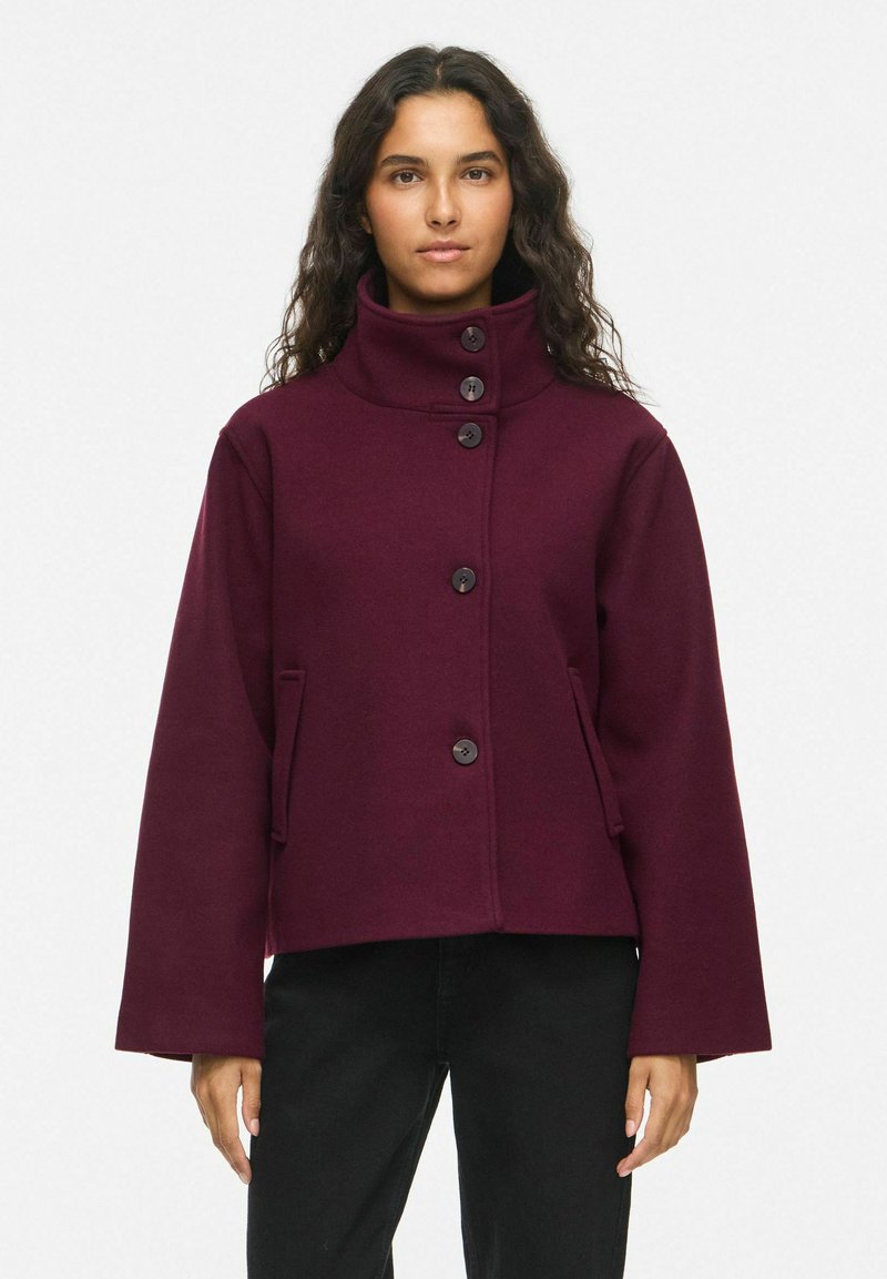 Femme aux longs cheveux bouclés portant une veste violette foncée à col montant boutonnée et un pantalon noir, debout sur un fond blanc.
