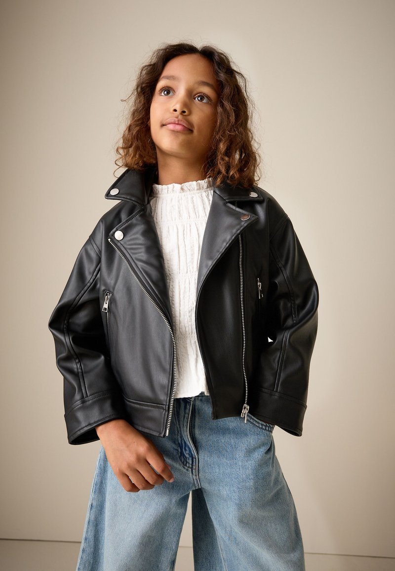 Niño con cabello rizado que lleva una chaqueta de cuero negra, blusa blanca texturizada y vaqueros azules holgados, de pie frente a un fondo beige liso.