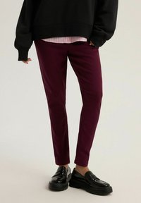 Osoba ubrana w ciemne burgundowe slim fit spodnie, czarne mokasyny, czarny oversize'owy sweter i różową koszulę w paski pod spodem.