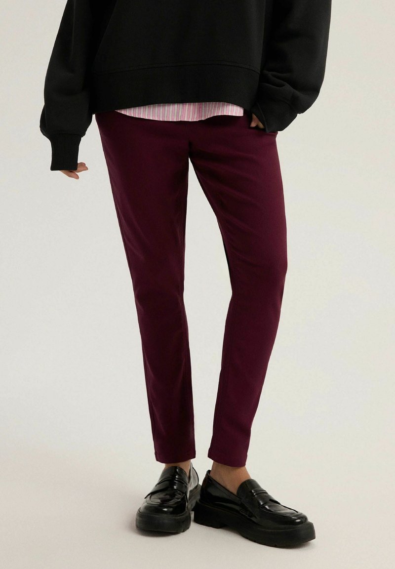 Osoba ubrana w ciemne burgundowe slim fit spodnie, czarne mokasyny, czarny oversize'owy sweter i różową koszulę w paski pod spodem.