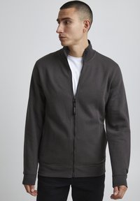 Felpa grigia con zip, realizzata in tessuto morbido, con colletto alto, tasche frontali e polsini a costine, abbinata a una t-shirt bianca sotto.