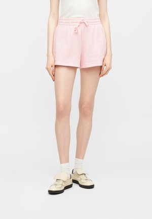 Γυναίκα που φοράει ροζ ανοιχτό σορτς με κορδόνι, λευκές κάλτσες και κρεμ αθλητικά παπούτσια, στέκεται μπροστά σε απλό φωτεινό φόντο.