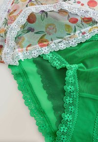 Sous-vêtements en dentelle verte transparente partiellement recouverts de tissu floral avec bordure en dentelle blanche sur un fond clair.