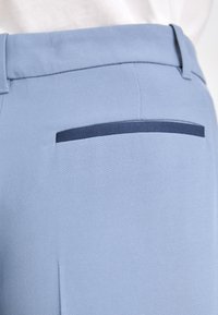 Pantalon bleu clair avec un tissu lisse et texturé, orné d'un accent en ruban marine sur la poche arrière. Coupe ajustée avec un design épuré.