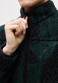 Το χέρι ενός ατόμου ανεβάζει το φερμουάρ σε μια ζακέτα από φλις με υφή, σε σκούρο πράσινο και μαύρο, με ψηλό γιακά.