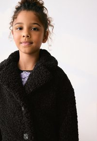 Manteau noir texturé et duveteux avec un large col et une fermeture à boutons, porté par-dessus un haut brillant à motifs avec un col rond.