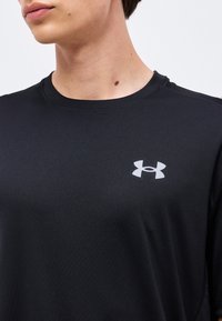 Νέος άνθρωπος φοράει μαύρο μπλουζάκι Under Armour, δείχνοντας λαιμό και μερικό πρόσωπο γυρισμένο προς τα πλάγια.