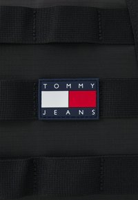 Tessuto nero con trama, dotato di cinghie orizzontali. Presenta una patch rettangolare blu navy con il nome del marchio "TOMMY JEANS" e loghi colorati.