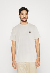 T-shirt in cotone beige chiaro con maniche corte, colletto tondo, caratterizzato da una piccola grafica multicolore sul lato sinistro del petto. Texture morbida.