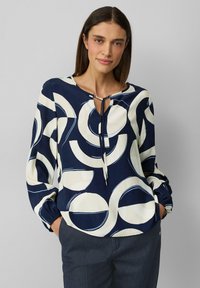Blouse marine avec de grands motifs circulaires crème et bleu clair, dotée d'un décolleté en forme de trou de clé avec un nœud, de manches longues et de poignets froncés.
