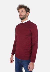 Maglione bordeaux a maglia con scollo rotondo e polsini a coste, abbinato a jeans di denim blu. Texture liscia, design aderente.