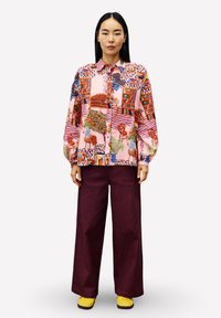 Eine Frau trägt ein farbenfrohes, gemustertes Hemd mit Knöpfen, dunkle weite Hosen und gelbe Schuhe und steht aufrecht vor einem einfarbigen Hintergrund.