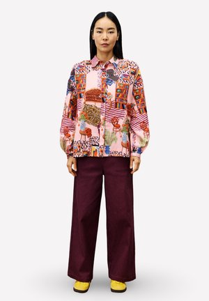 Femme portant une chemise à boutons à motifs colorés, un pantalon large foncé et des chaussures jaunes, se tenant droite contre un fond uni.