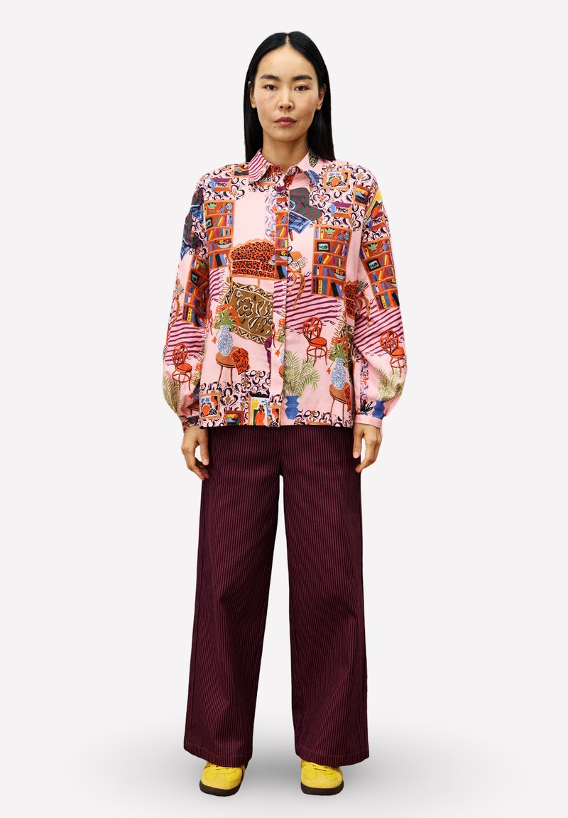 Eine Frau trägt ein farbenfrohes, gemustertes Hemd mit Knöpfen, dunkle weite Hosen und gelbe Schuhe und steht aufrecht vor einem einfarbigen Hintergrund.