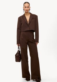 Femme portant un blazer court marron chocolat et un pantalon à jambes larges, tenant un sac à main à sequins, avec des boucles d'oreilles argentées pendantes et des escarpins à nœud.
