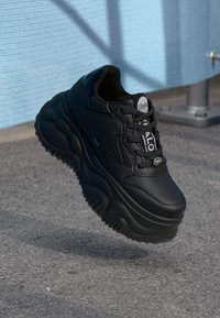 Svart sneaker med en tykk, bølgete gummisåle, glatt skinnoppe, rund logoetikett og teksturerte snører med hvit aksent.