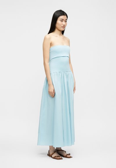 Γυναίκα που στέκεται με ανοιχτό μπλε φόρεμα maxi χωρίς τιράντες και καφέ επίπεδα σανδάλια, μπροστά σε απλό λευκό φόντο.