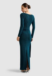 Dlhé tyrkysové šaty s dlhými rukávmi, trblietavým materiálom, zipsom na chrbte, bočným rozparkom a záhybmi v páse. Strieborné vysoké opätky.