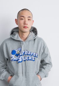 Haut à capuche gris avec un graphique à l'avant affichant "Carhartt Bucks" en bleu, mettant en avant un personnage de dessin animé et des éléments de basket-ball.
