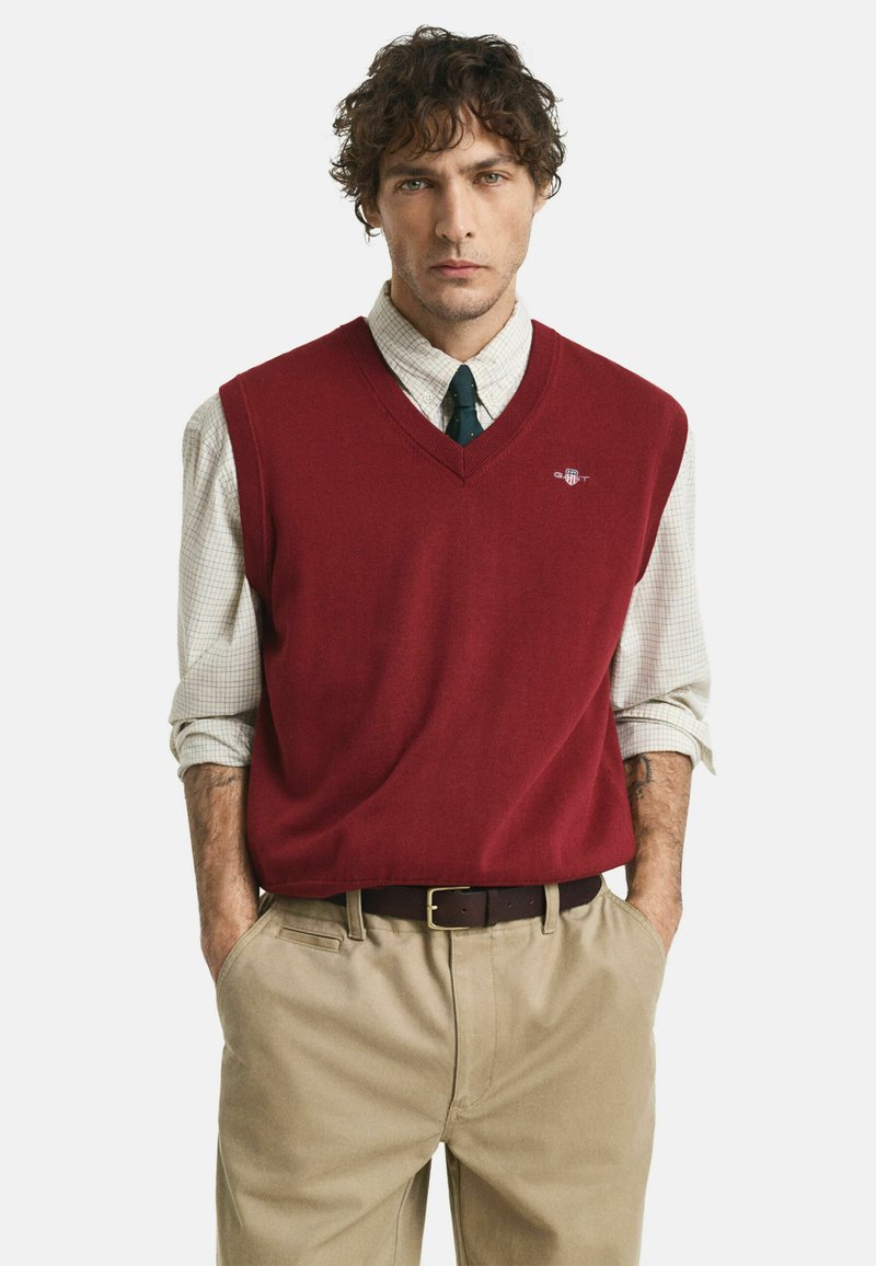 Gilet rosso lavorato a maglia con scollo a V indossato sopra una camicia a quadri, abbinato a una cravatta scura e pantaloni beige, con una cintura marrone.