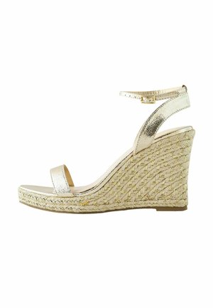 Sandał na koturnie z plecioną jucie platformą, metalicznym złotym paskiem wokół kostki oraz odkrytymi palcami na białym tle.
