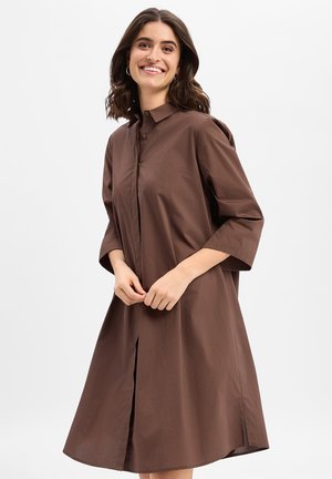 Lächelnde Frau mit welligem dunklem Haar, die ein locker sitzendes braunes Hemdkleid mit Dreiviertelärmeln vor einem schlichten weißen Hintergrund trägt.