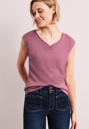 Smilende kvinde med kort blondt hår iført en ærmeløs malva top og mørkeblå jeans mod en beige baggrund.