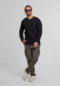 Schwarzes Langarm-Henley-shirt mit Knopfleiste, kombiniert mit grünen Cargo-Hosen mit Seitentaschen und weißen Turnschuhen.