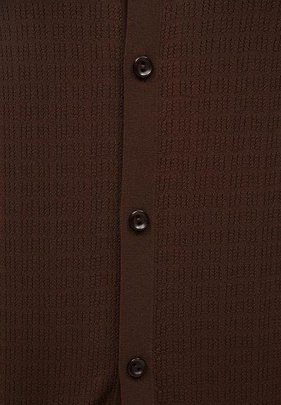 Κοντινή λήψη καφέ υφασμάτινης υφής με μια κάθετη σειρά από τρία σκούρα καφέ κουμπιά ραμμένα σε μια μονή ζώνη.