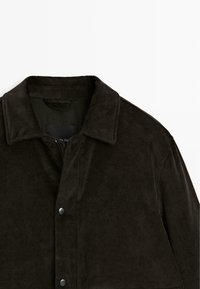 Giacca di velluto a coste marrone scuro con colletto, bottoni a pressione e tessuto testurizzato. Presenta un taglio ampio e un design minimalista.