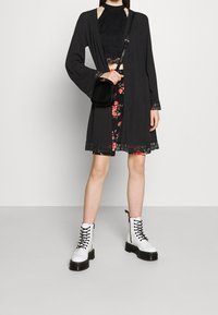 Kvinna i svart spetsblus, svart kort kofta med spetsdetaljer, blommig kjol, vita snörstövlar och håller en svart crossbody-väska.