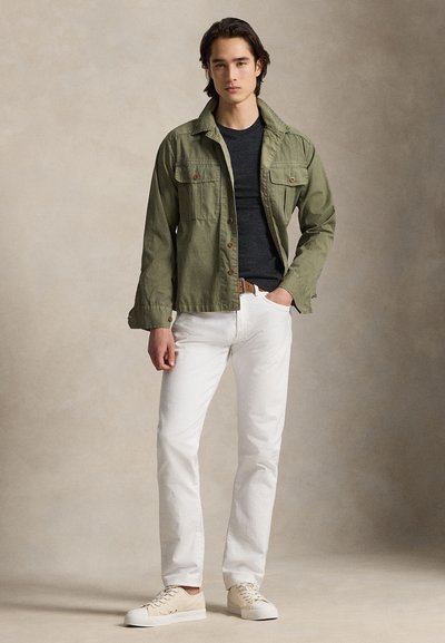 Olivengrønn bomullsjakke med to frontlommer, svart genser med rund hals, hvite bukser og beige joggesko. Glatt tekstur og tettsittende design.