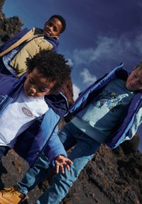 Três crianças com casacos coloridos: um casaco acolchoado azul escuro, um colete bege e uma sweat azul clara. O fundo apresenta um terreno rochoso.