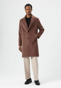 Bruine mantel van een wolmix met een kentkraag, enkele knoopsluiting, zijzakken en een gladde textuur; het model combineert het met lichte broek en zwarte schoenen.