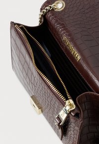 Hnedá kabelka z kože so vzorom krokodíla s zlatým reťazovým popruhom, zlatým zipsom a logom "Steve Madden" na klapke.