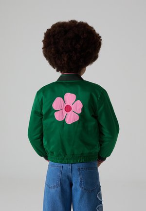 Enfant aux cheveux bouclés portant une veste verte avec un grand motif de fleur rose dans le dos et un jean bleu.