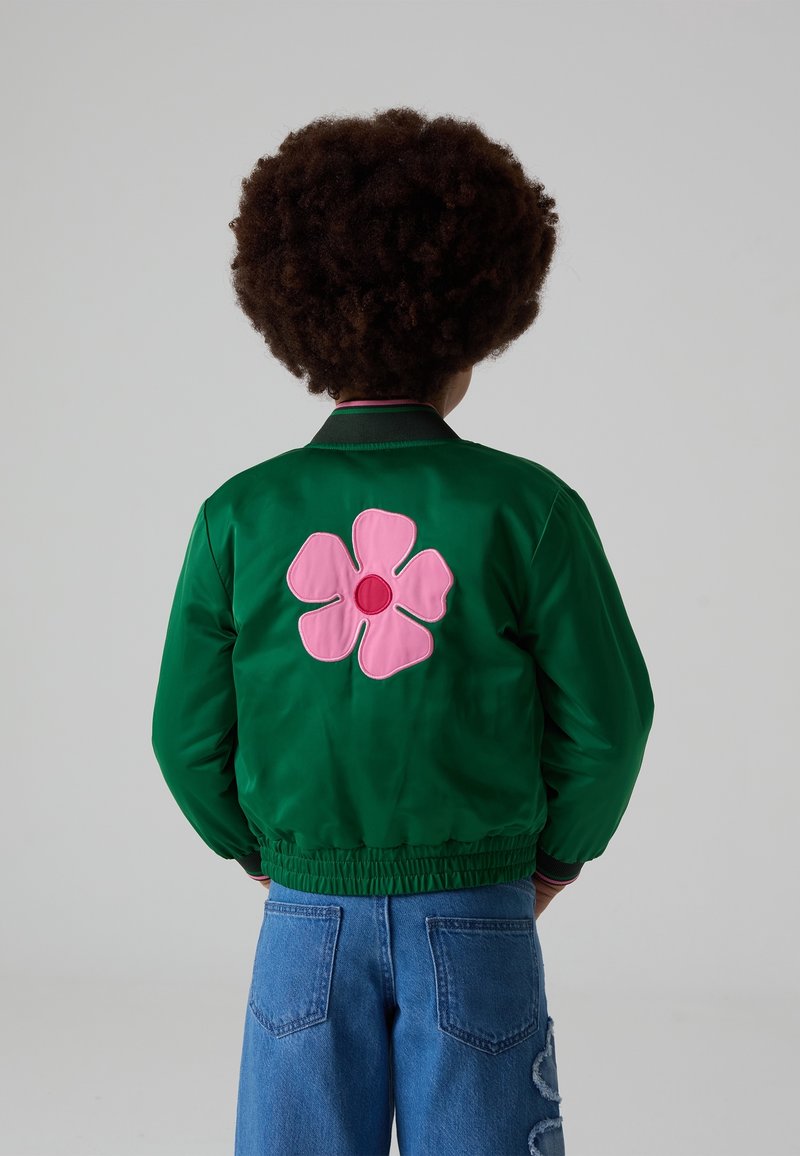Enfant aux cheveux bouclés portant une veste verte avec un grand motif de fleur rose dans le dos et un jean bleu.