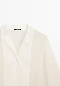 Chemise légère, écrue, à manches longues, avec un design en tissage ouvert texturé et un col classique. Présente des détails de boutons subtils.